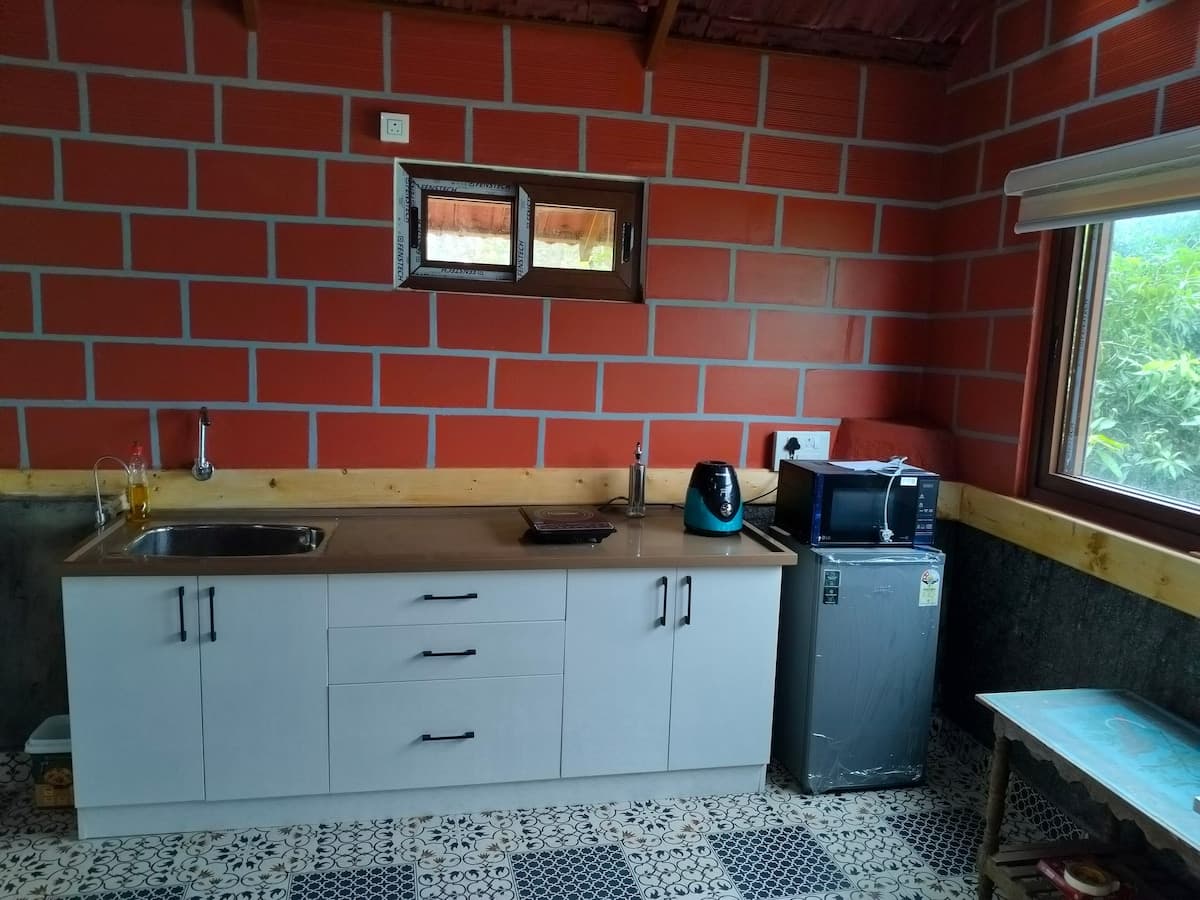 The kitchen counter along a brick wall: a sink, a kettle, a microwave and a small fridge, over white cabinets, on a patterned tile floor.