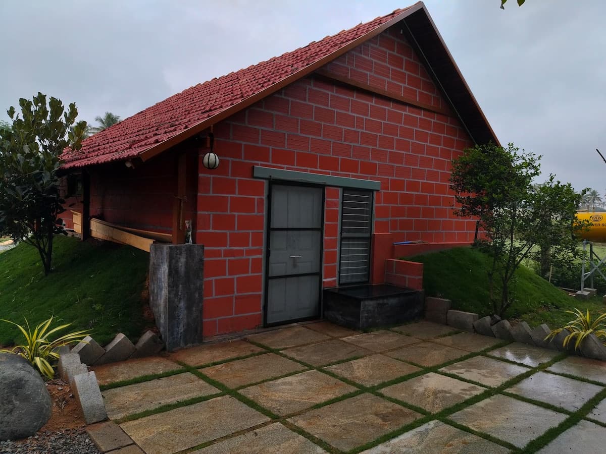 The gable end of the house after rain: red brick under a Mangalore tile roof, a grey steel door and a louvred window, wet flagstone paving with grass in the joints, and grassed banks rising to the walls on both sides.