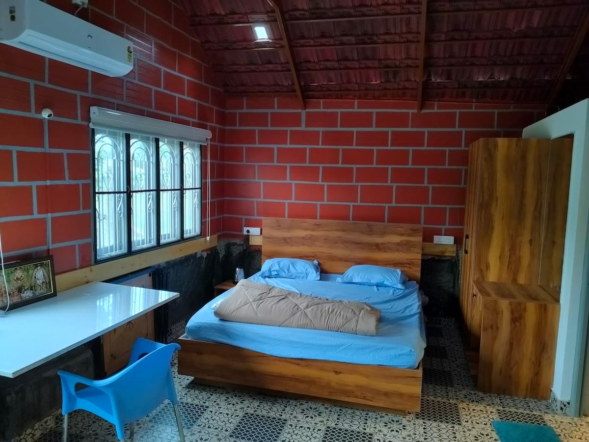 Inside the single room: a bed made up in blue against a bare brick wall, a white table and a blue plastic chair under the grilled window, an air conditioner high on the wall, and the underside of the tile roof carried on wooden rafters.