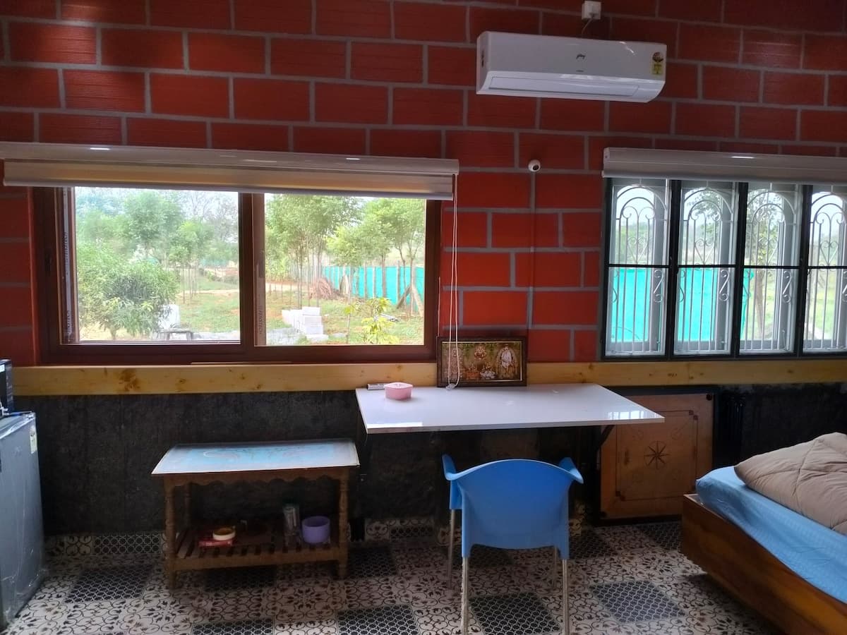 The desk under the windows: a white worktop against the brick wall, a chair drawn up to it, an air conditioner above, and the orchard and boundary fence bright through the glass on both sides.