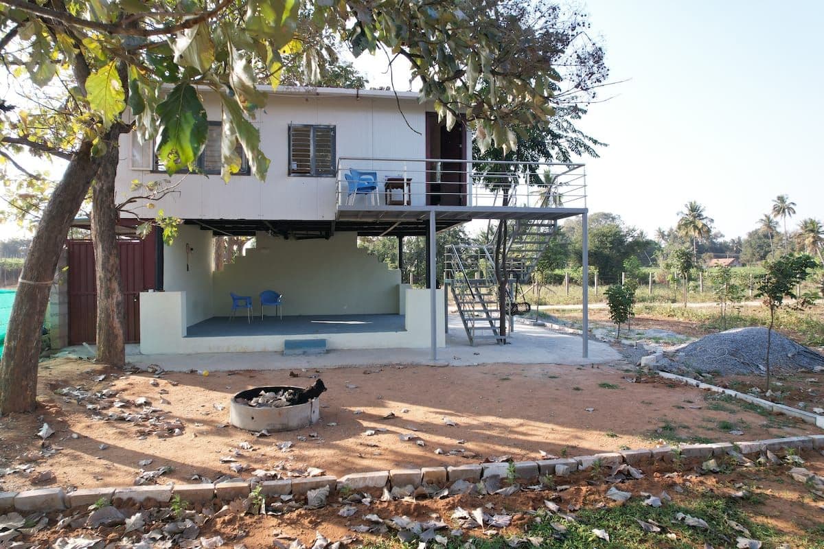 The tree house from the front with its open shaded ground floor, the steel stair up to the deck, and a circular fire pit set in the swept earth in front of it.