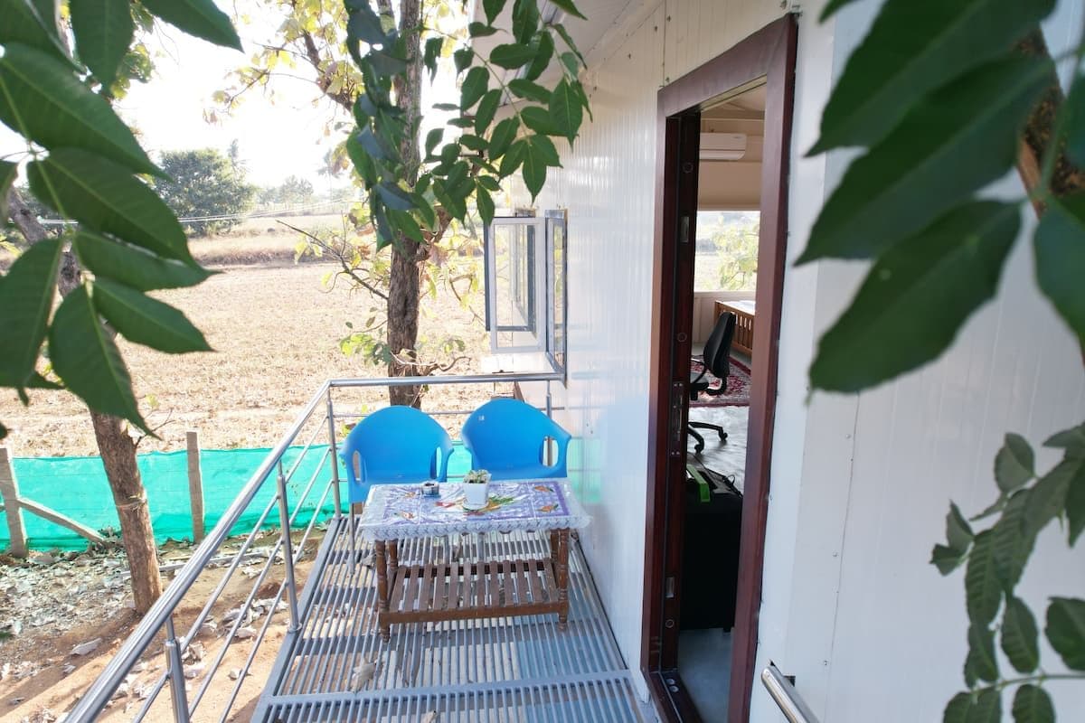 The upper deck: a wooden table and blue chairs on steel grating outside an open door, with tree canopy at the railing and a field beyond it.