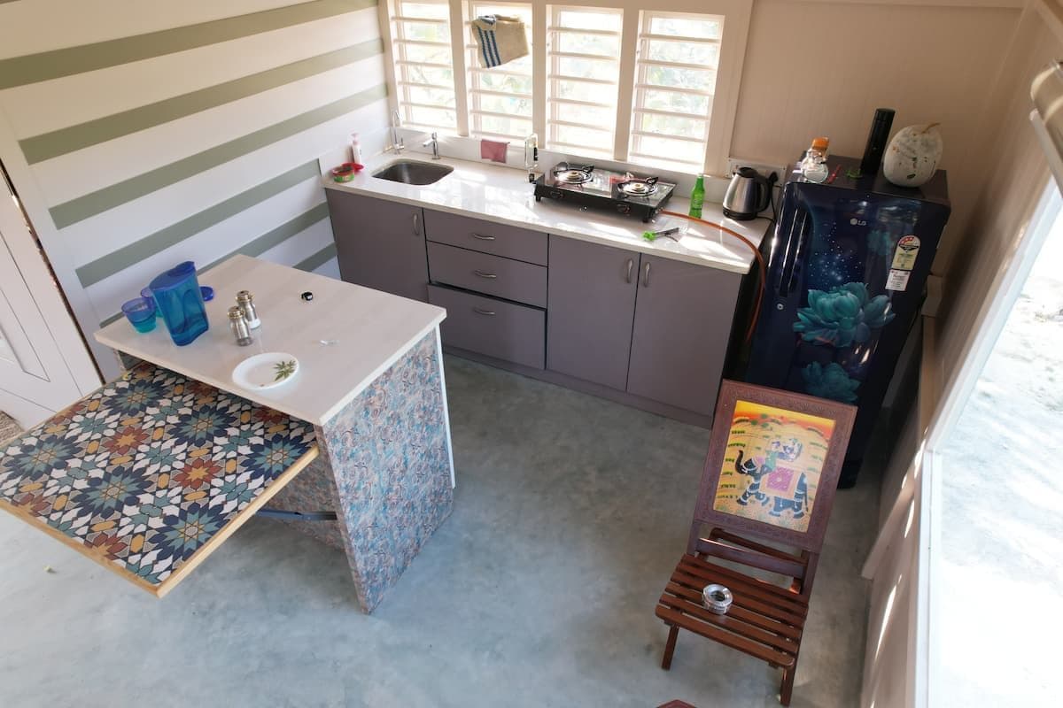 The kitchen end of the upper room from above: a drop-leaf table faced in patterned tile, a counter with a sink and a gas ring under the louvred windows, and a fridge in the corner.