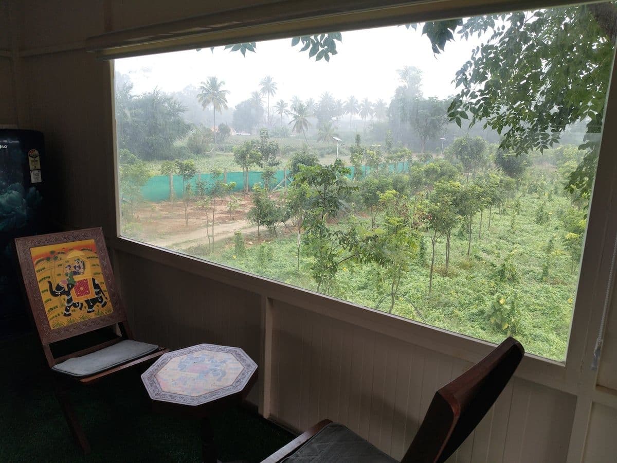 The window corner of the upper room in the early morning: two carved folding chairs and a small painted octagonal table, with the orchard and the boundary fence soft in mist through the glass.