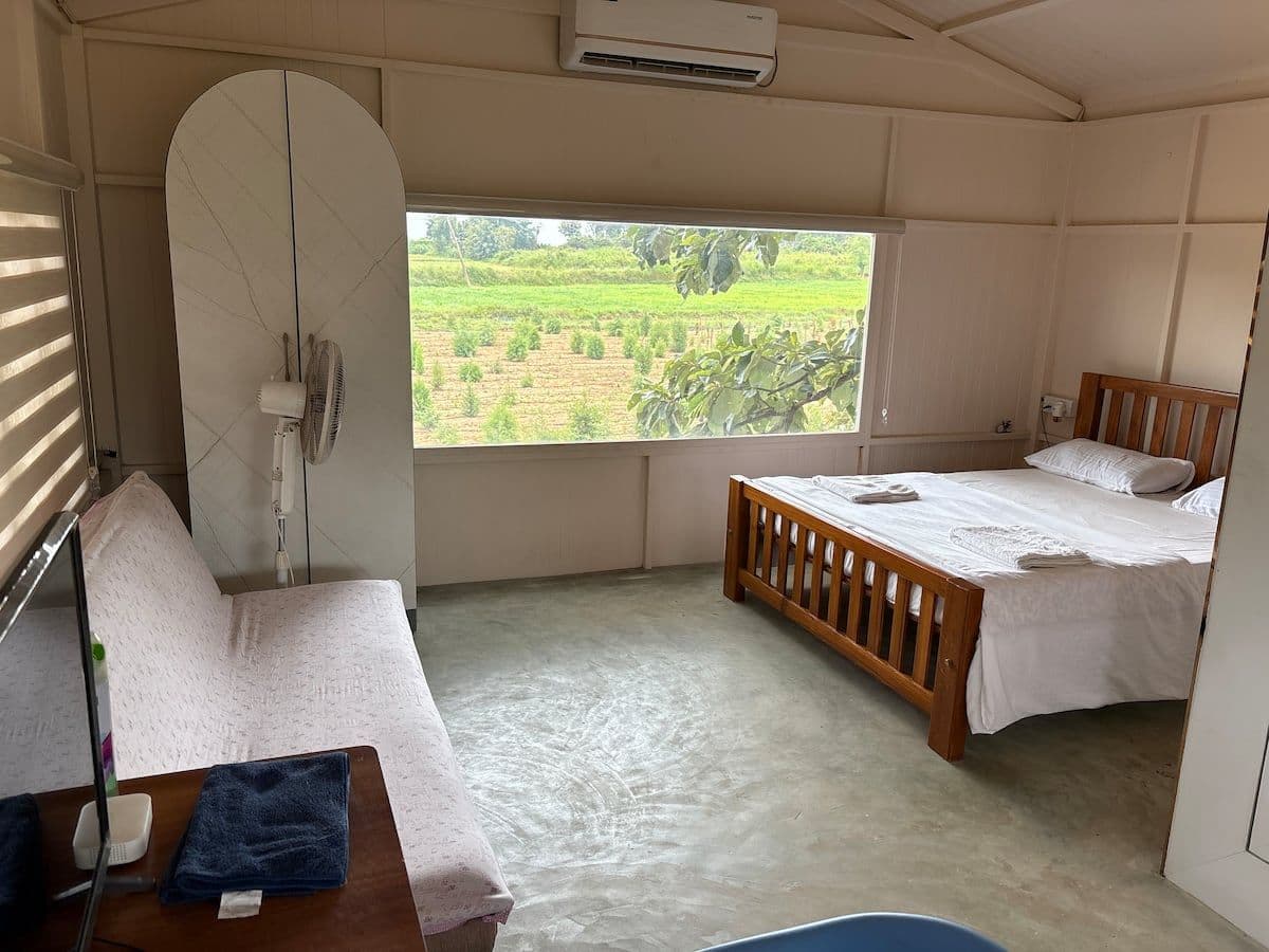The bedroom: a wooden-framed double bed made up in white with towels folded on it, a curved wardrobe, an air conditioner, and a wide window onto a green field.